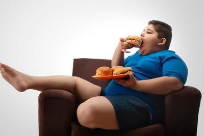 Obese young boy eating burger sitting on sofa