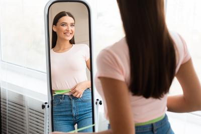 Young woman smiling in the mirror with tape measure in hip.