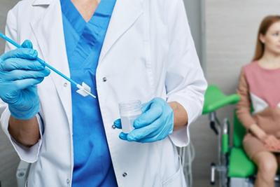 Close-up of gynecologist holding Pap smear test vial with woman in background.