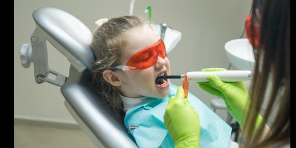 A young girl sits in the dentist’s chair and has her teeth examined by a female dentist.