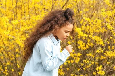 Young-girl-in-a-blue-shirt-coughing-outdoors-near-bright-yellow-flowers.