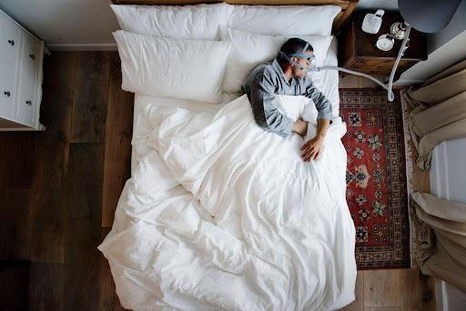 A man lies sleeping in a bed. He is wearing a CPAP machine. The sheets, pillows, and blanket are all white in color. There is a window to the man’s left.