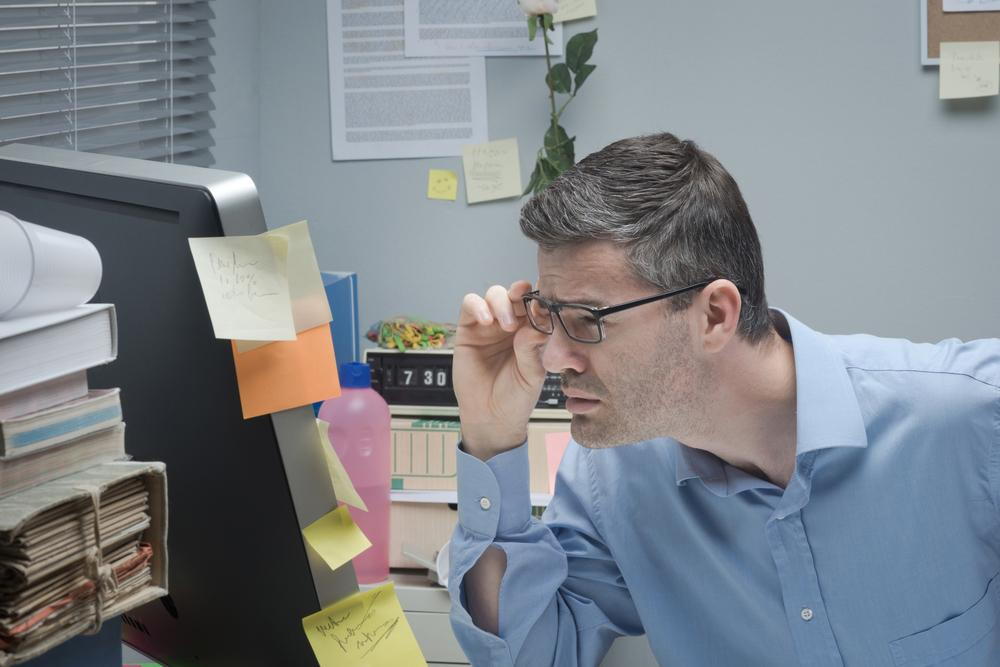 man at his desk squinting to see his computer screen due to straining his eyes at work