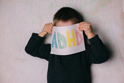 Child holding a sheet of paper with the word ADHD written in crayons covering his face.