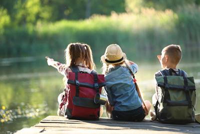 Three kids with backpacks sit on a dock by the lake, enjoying the natural scenery.