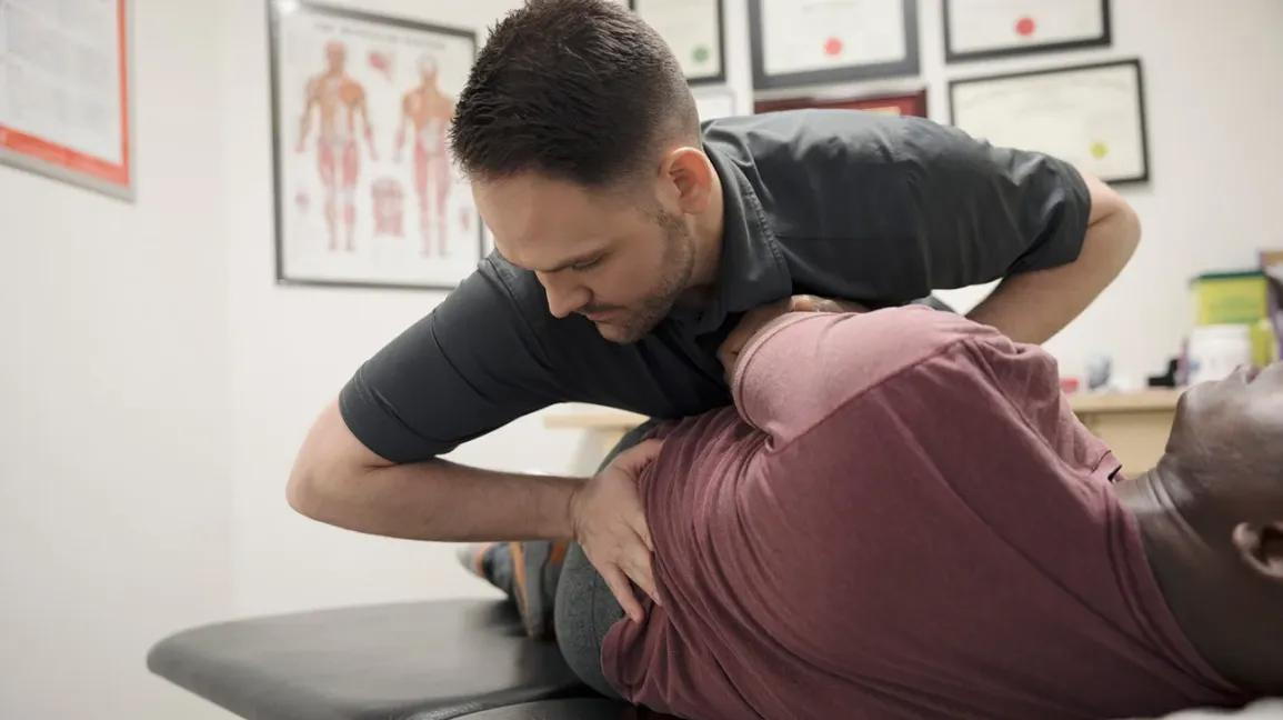 A chiropractor does a spinal manipulation on a patient who is lying on a chiropractic table in his office.