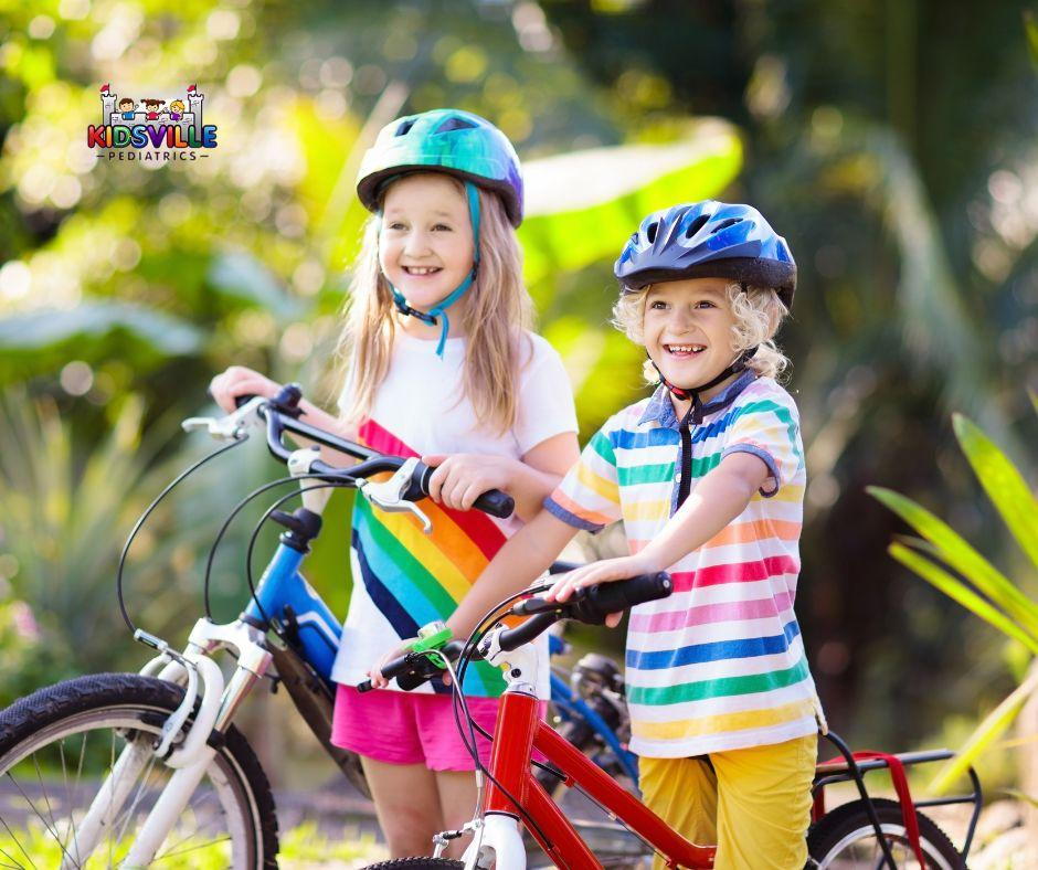 alt= Two children wearing helmets and vibrant clothes stand next to their bicycles outdoors. The logo in the top left corner reads “Kidsville Pediatrics,” emphasizing a commitment to child health.