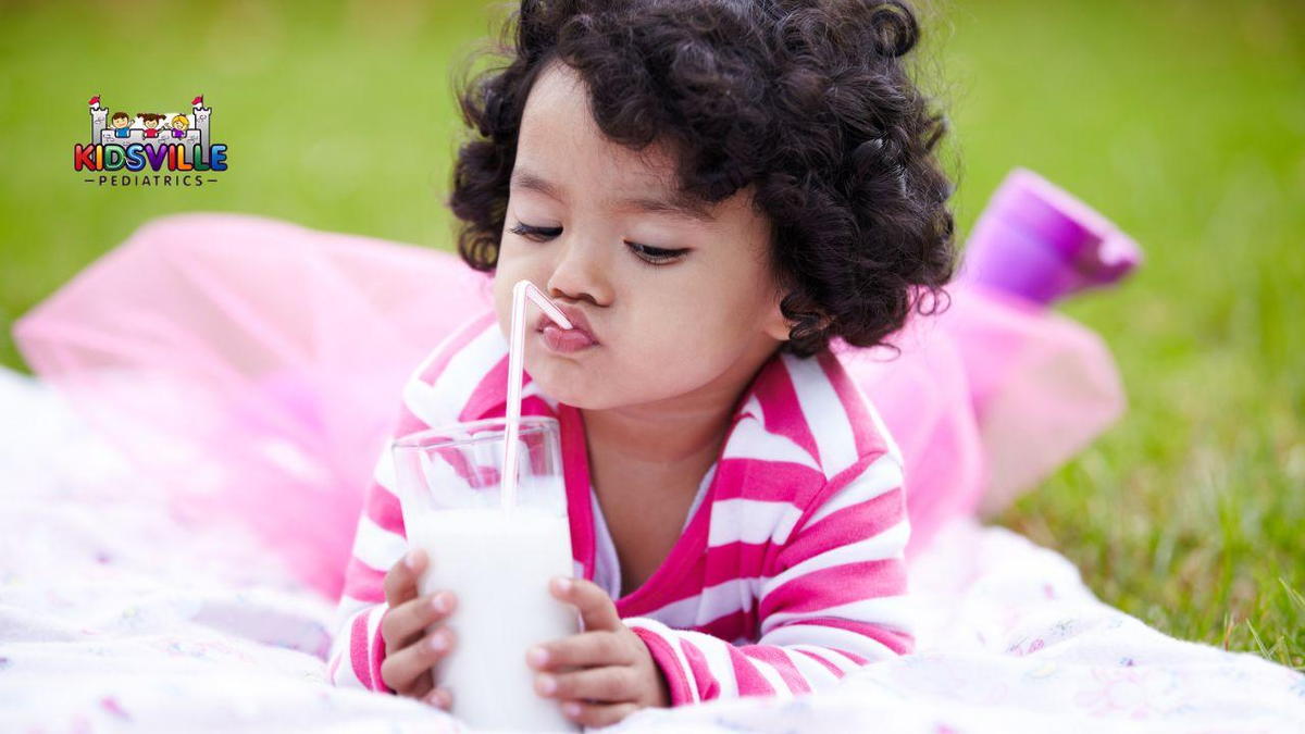 A baby girl lies on the grass, smiling, with a glass of milk beside her, enjoying a sunny day outdoors.