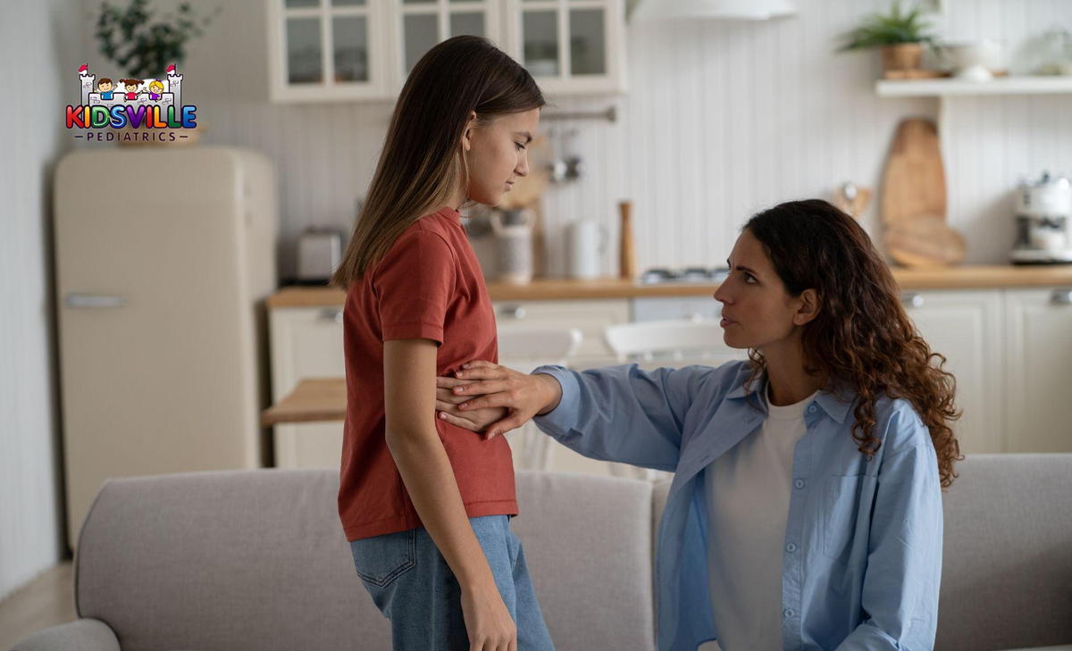 A woman looks concerned as a young woman clutches her lower right abdomen in front of her.