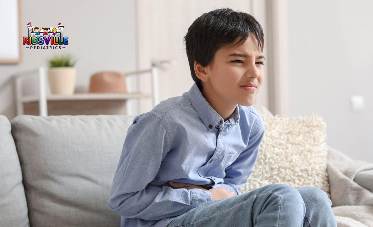 A young boy sits on a couch, holding his stomach in discomfort, showing signs of pain on his face.