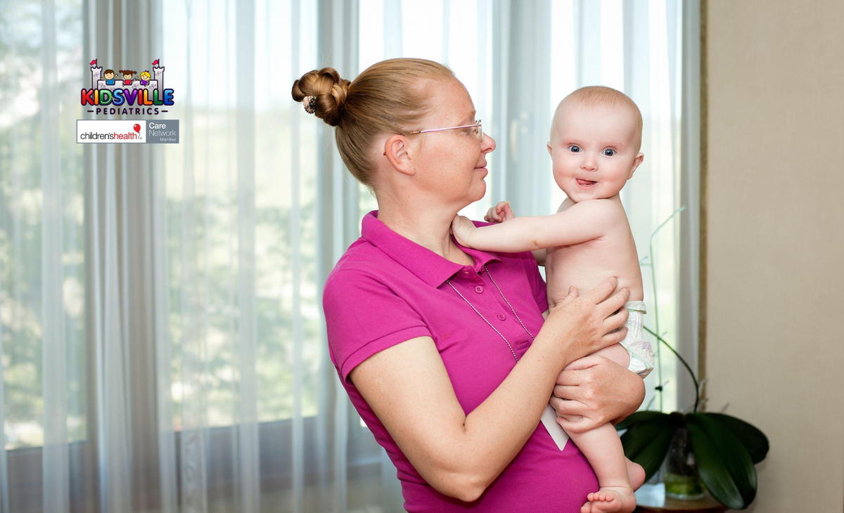 A woman gently cradles a baby in her arms, standing in front of a bright window, showcasing a moment of tenderness.