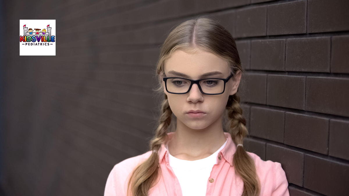 A young woman with glasses is standing, pensively, beside a brick wall.