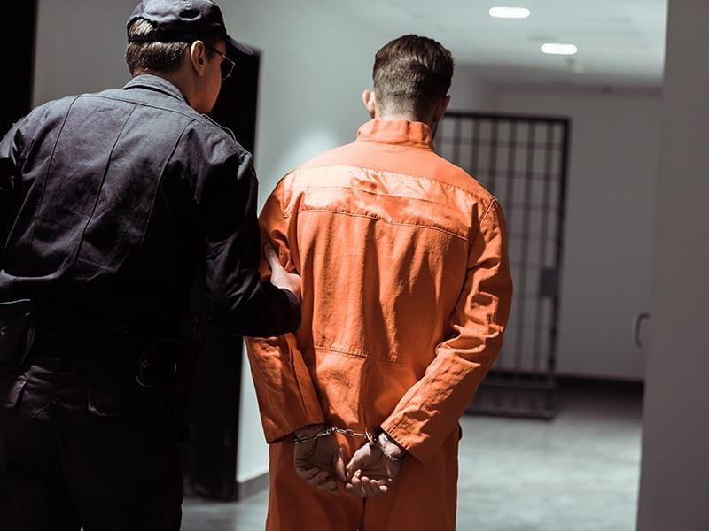 A man in handcuffs and prison orange jumpsuit being escorted to a jail cell.